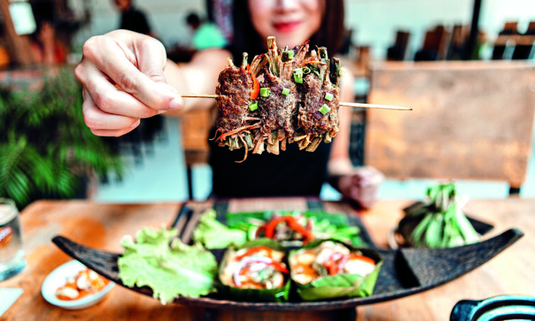 Close up shot of young Asian woman having freshly made authentic Thai food in a Thai restaurant - BBQ Beef Skewers, Chicken Green Curry, Seafood Tom Yum Goong soup and Sticky Rice. With a glass iced water. Local street food stall. Authentic Thai street food theme. Bangkok, Thailand.